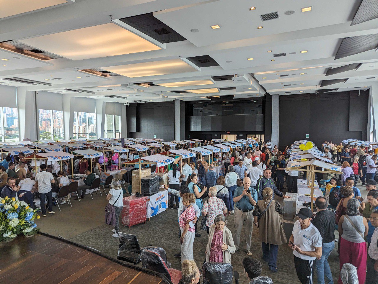 Import Resale Cross-border EC ist eine groß angelegte Veranstaltung in Innenräumen, bei der eine große Anzahl von Menschen um einen Tisch mit verschiedenen ausgestellten Gegenständen in einem Raum mit großen Fenstern und Blick auf die Stadt steht und sich unterhält.